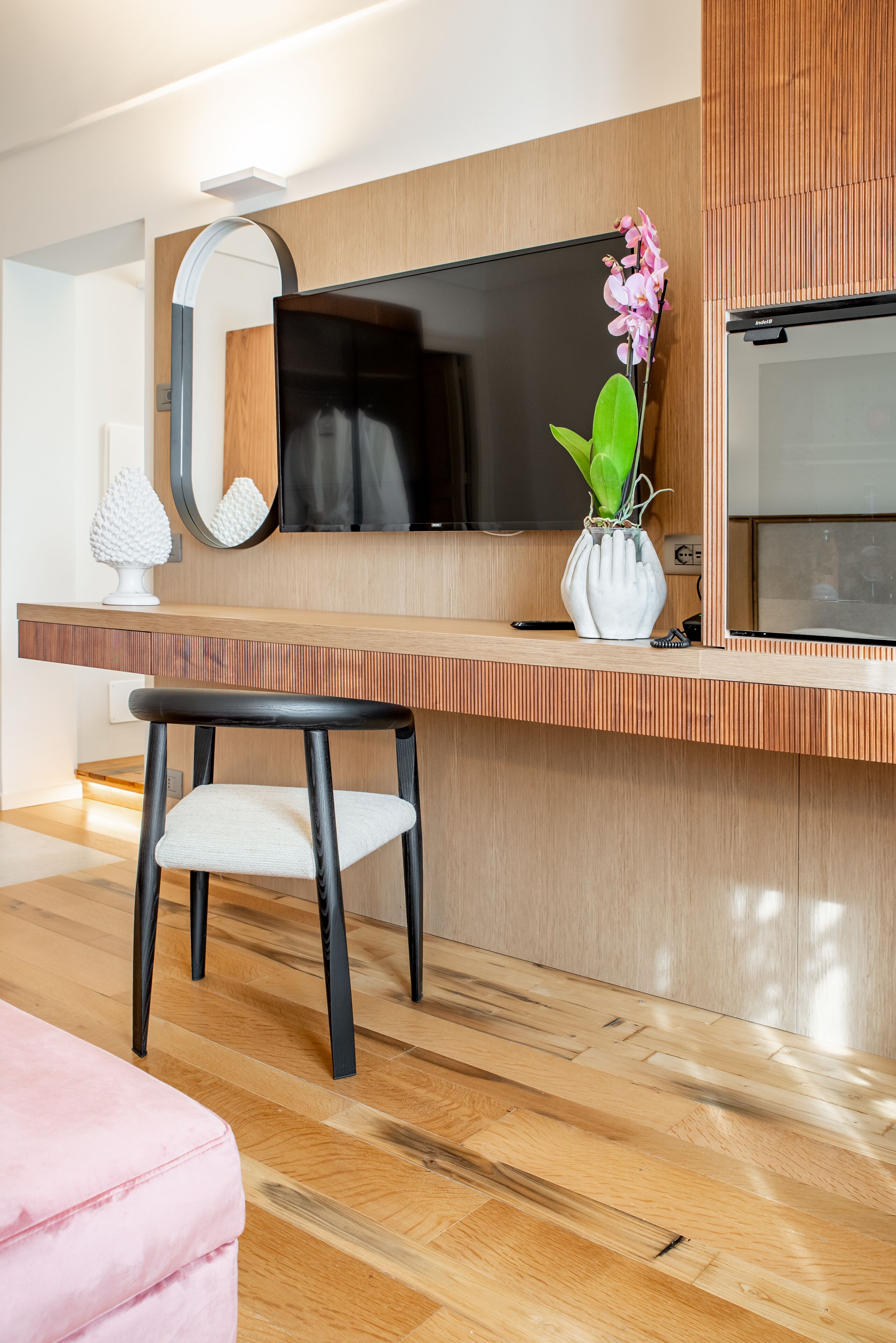 Una stanza moderna con pavimento in legno, un divano rosa, una sedia bianca, una TV, uno specchio e un vaso con una pianta.