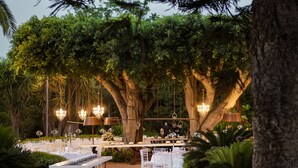 Un espacio para eventos al aire libre con mesas y sillas dispuestas bajo árboles grandes, rodeado de follaje.