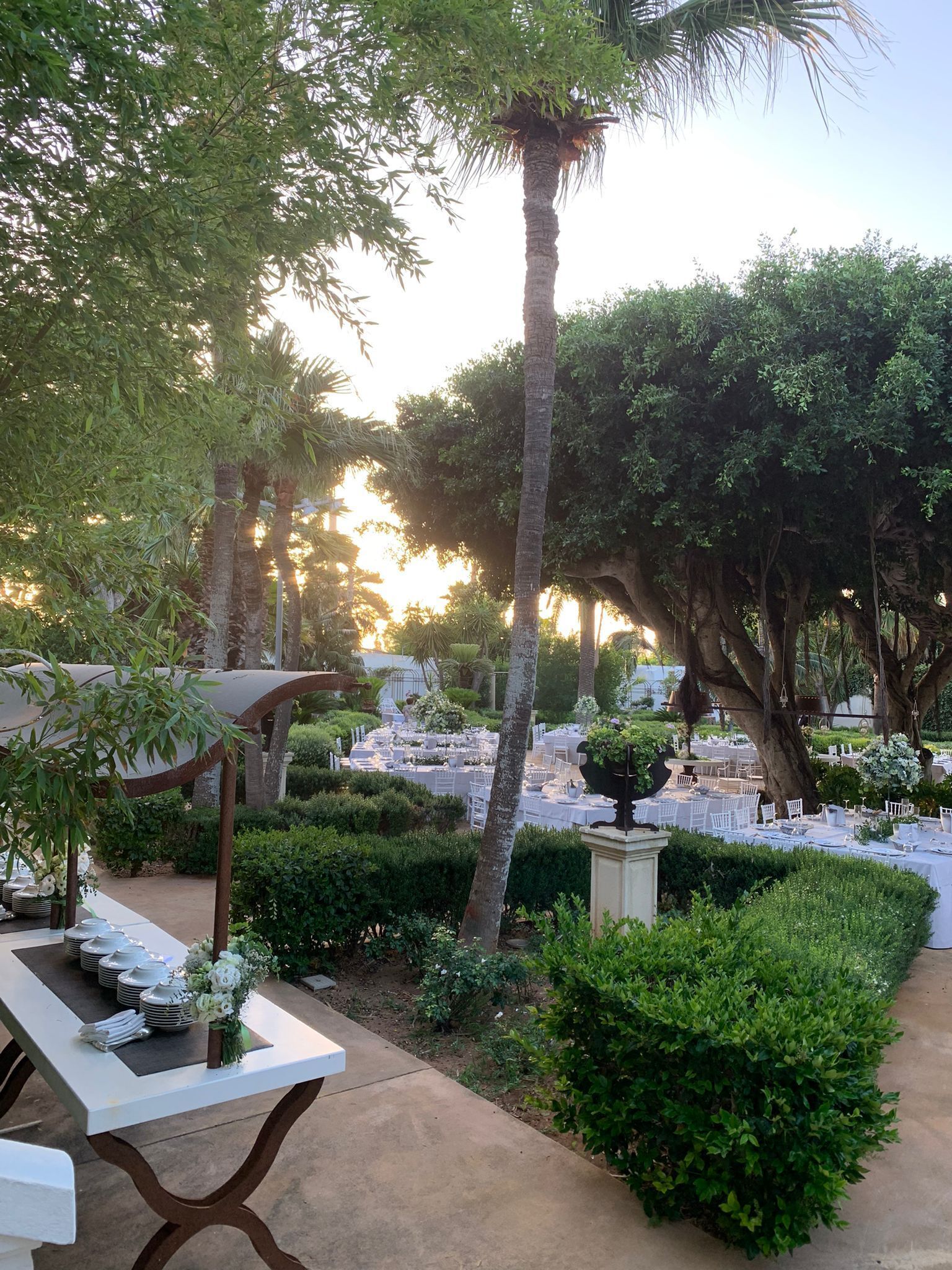 Un espace extérieur aménagé en jardin, avec des tables et des chaises disposées pour les repas, entouré d’une végétation luxuriante et de palmiers.