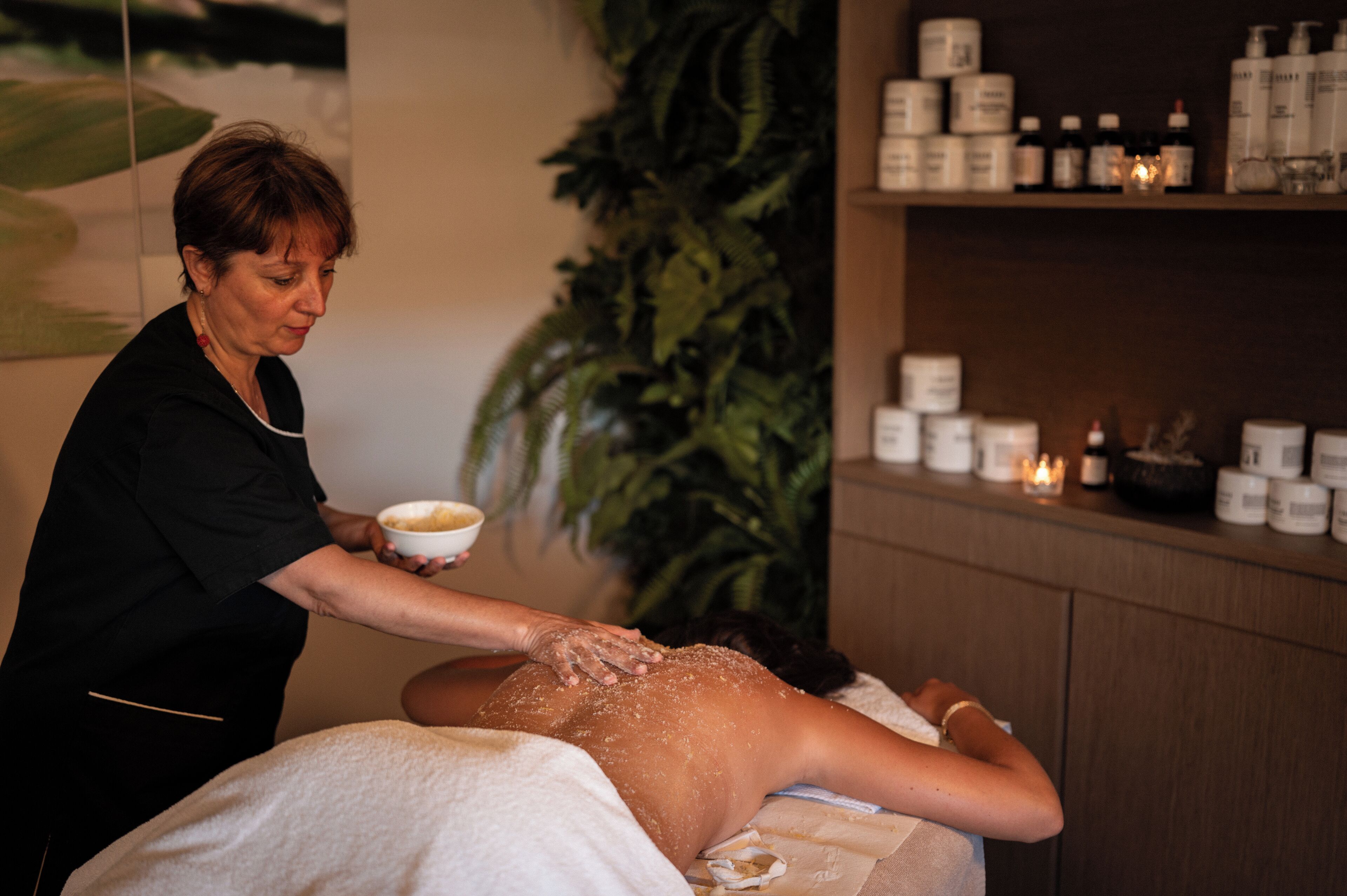 A person receiving a massage in a room with a large plant, shelves with bottles, and a candle.