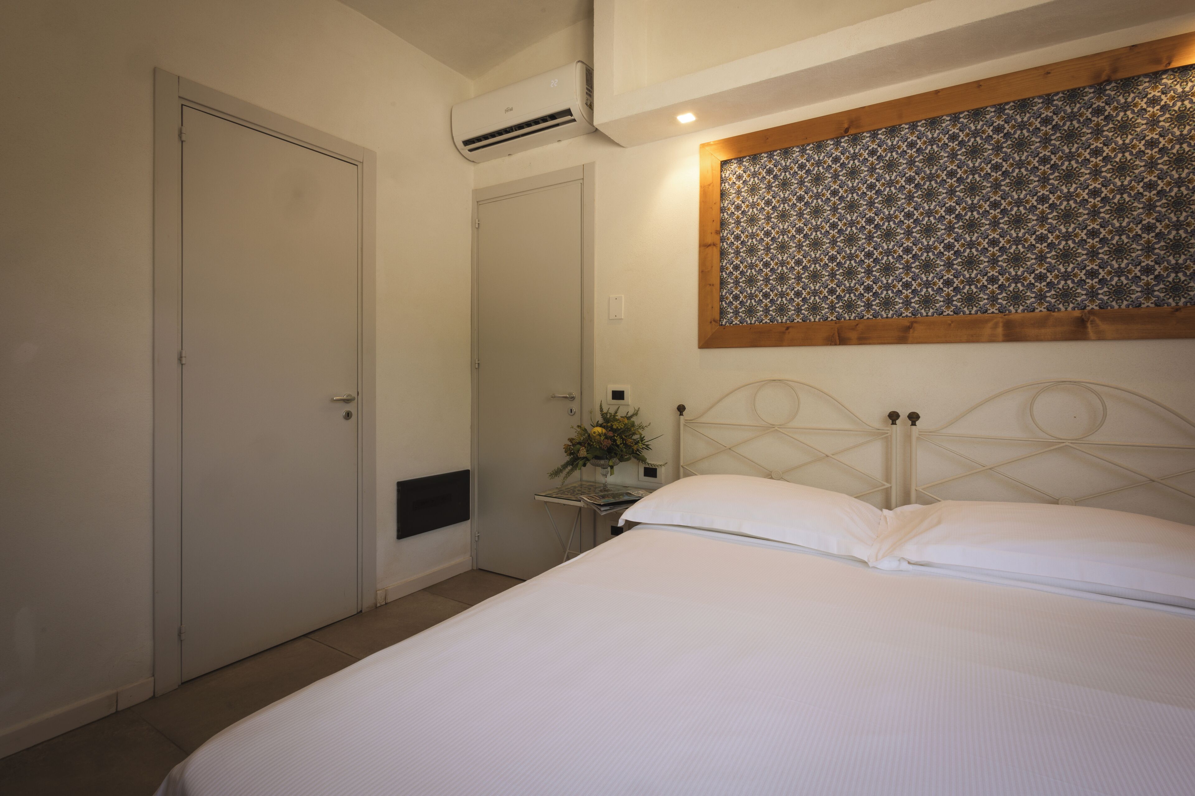 A bedroom with a bed, a door, an air conditioning unit, a small table with a vase, and a patterned wall above the bed.