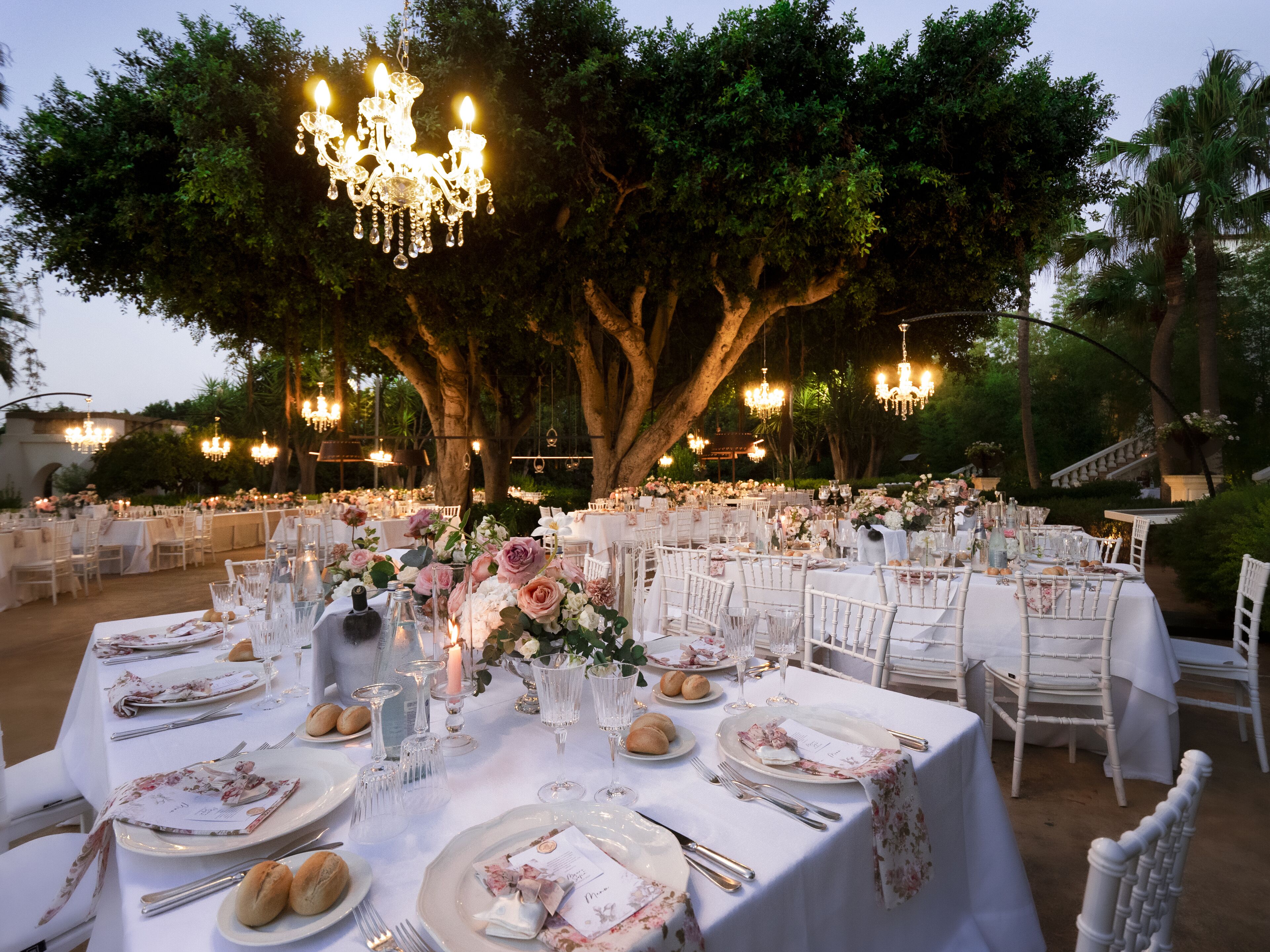 An outdoor banquet hall with tables set for a formal event, surrounded by trees and illuminated by chandeliers.
