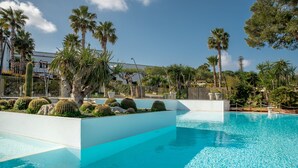 Una piscina con agua azul cristalina, rodeada de palmeras y cactus, con un edificio blanco al fondo.