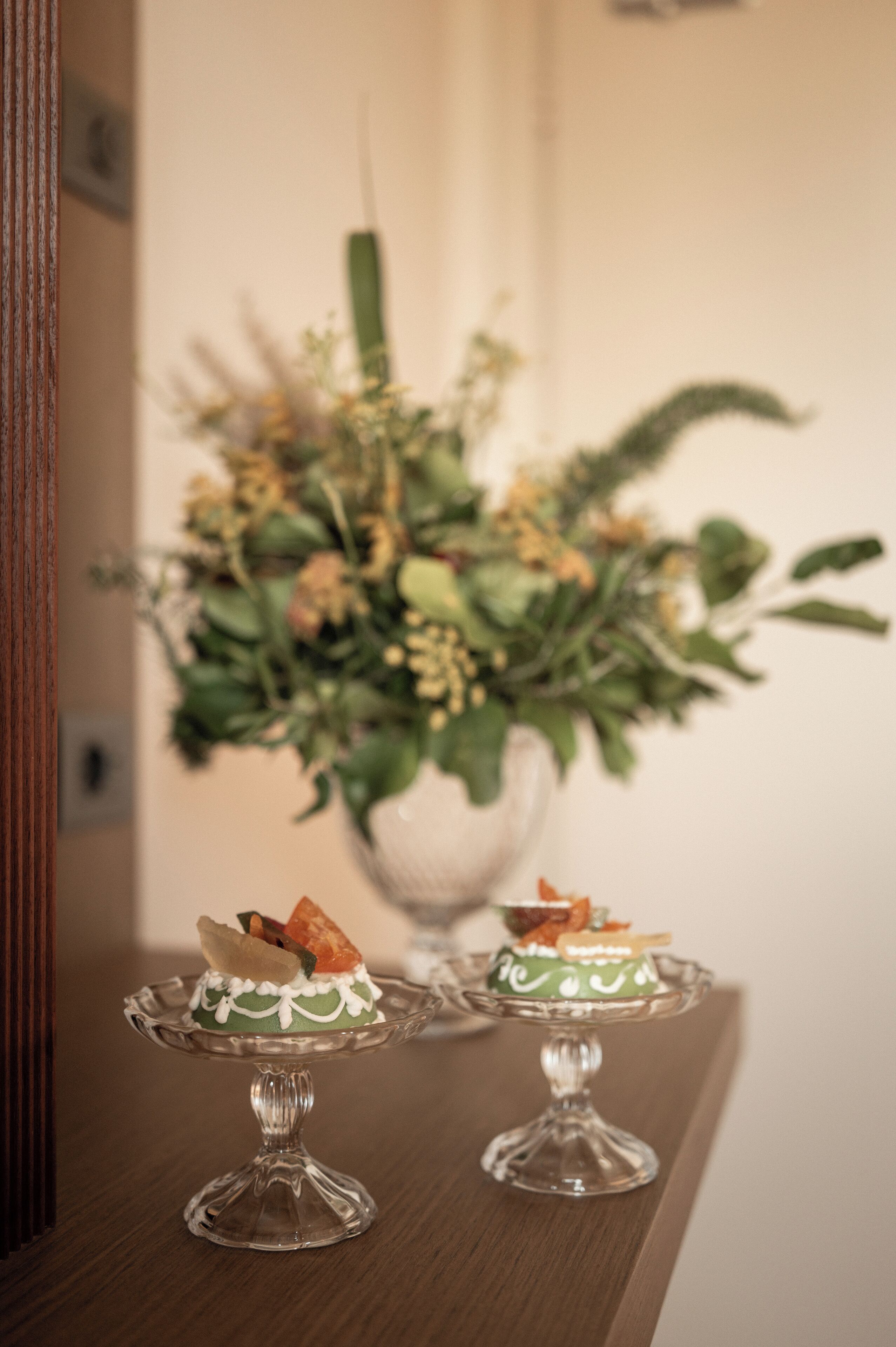 Two glass dessert stands on a wooden table with a blurred floral arrangement in the background.
