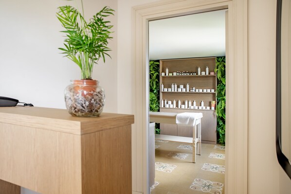 A reception area with a wooden desk, a potted plant, and a mirror reflecting a shelf with toiletries. - Villa Favorita (Marsala)
