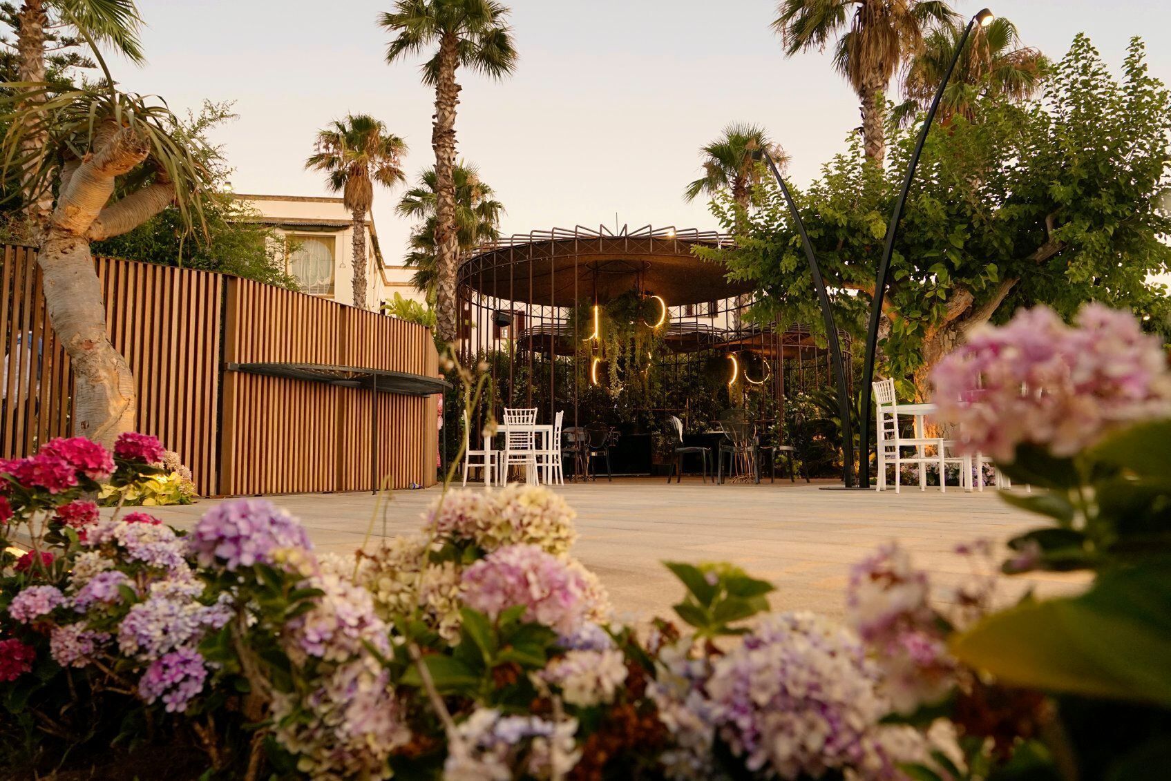 An outdoor seating area with tables and chairs, surrounded by palm trees and flowering plants.