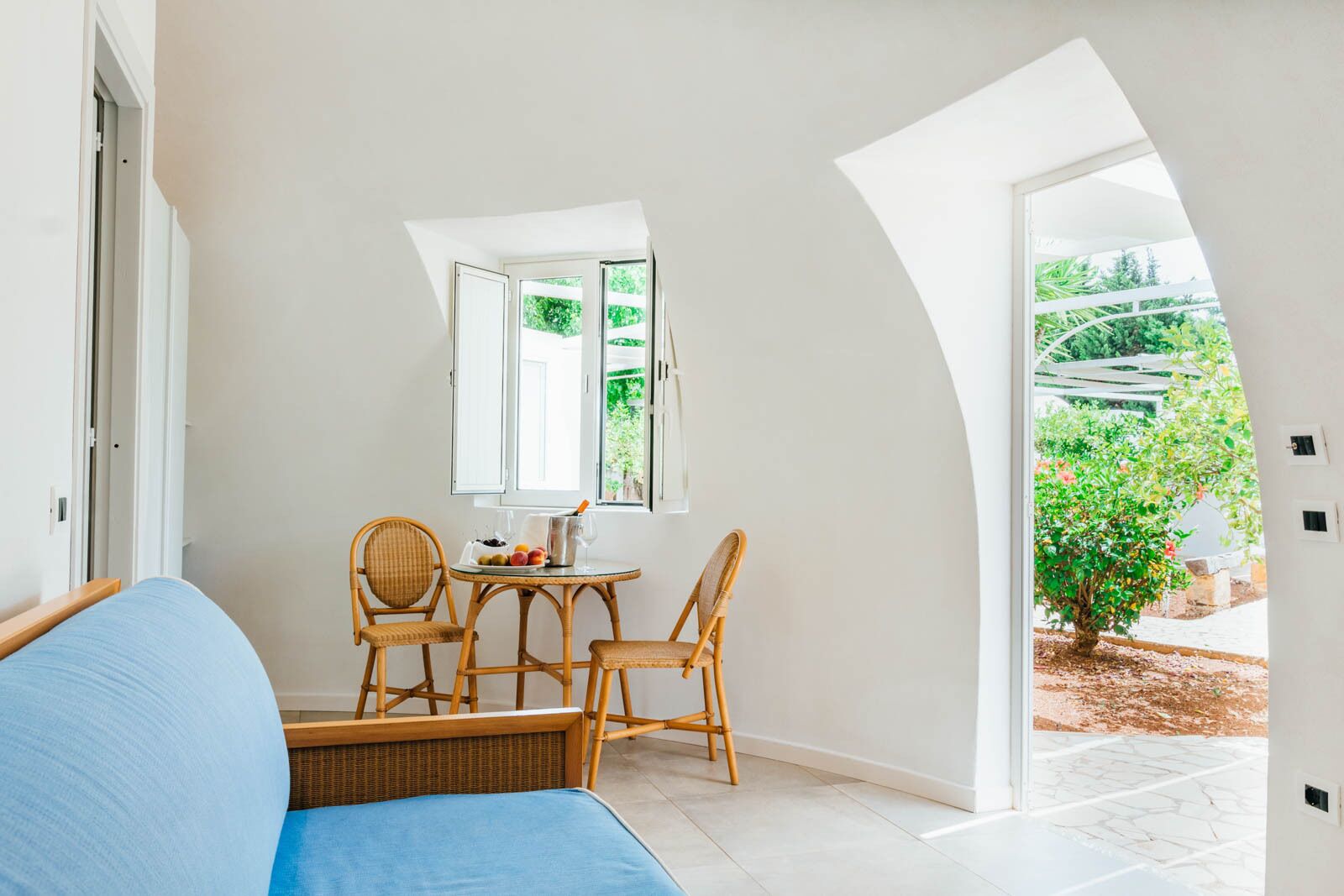 A room with a bed, a table with chairs, and a window with a view of greenery.