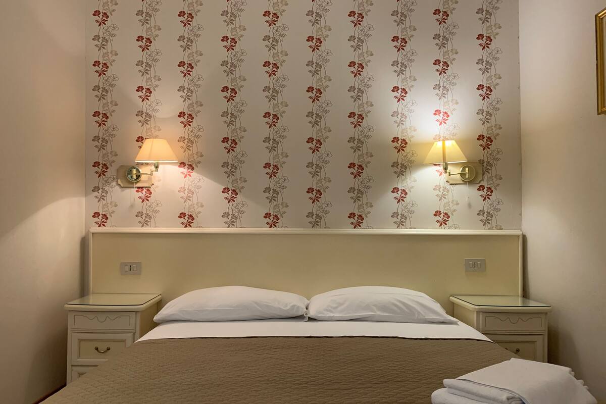 Een net opgemaakt bed met een bruine sprei, twee bedlampen en behang met een bloemenpatroon.