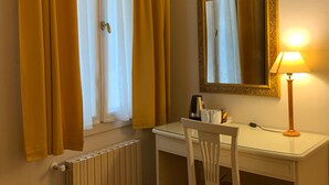 Una habitación pequeña y bien iluminada, con un escritorio blanco, una silla, un radiador, una ventana con cortinas amarillas y un espejo con una lámpara sobre el escritorio.