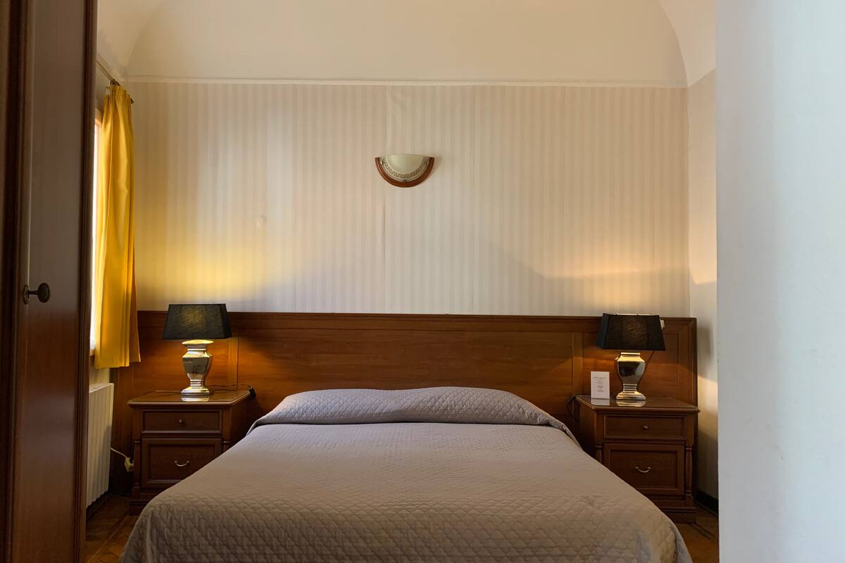 Een hotelkamer met een groot bed, twee nachtkastjes met lampen, een houten hoofdeinde en een raam met gordijnen.