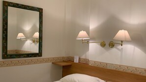 Habitación de hotel con una cama, ventilador de techo, espejo fijado a la pared y dos apliques con lámparas.