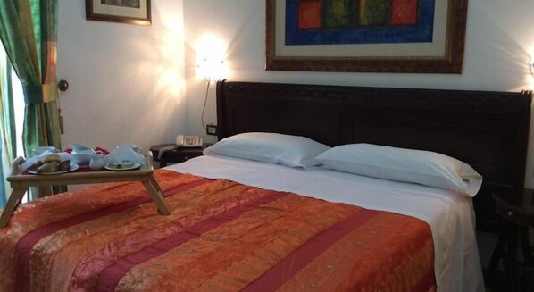 Una habitación de hotel con una cama, una mesita de noche con una bandeja de pasteles, una lámpara y un cuadro en la pared.