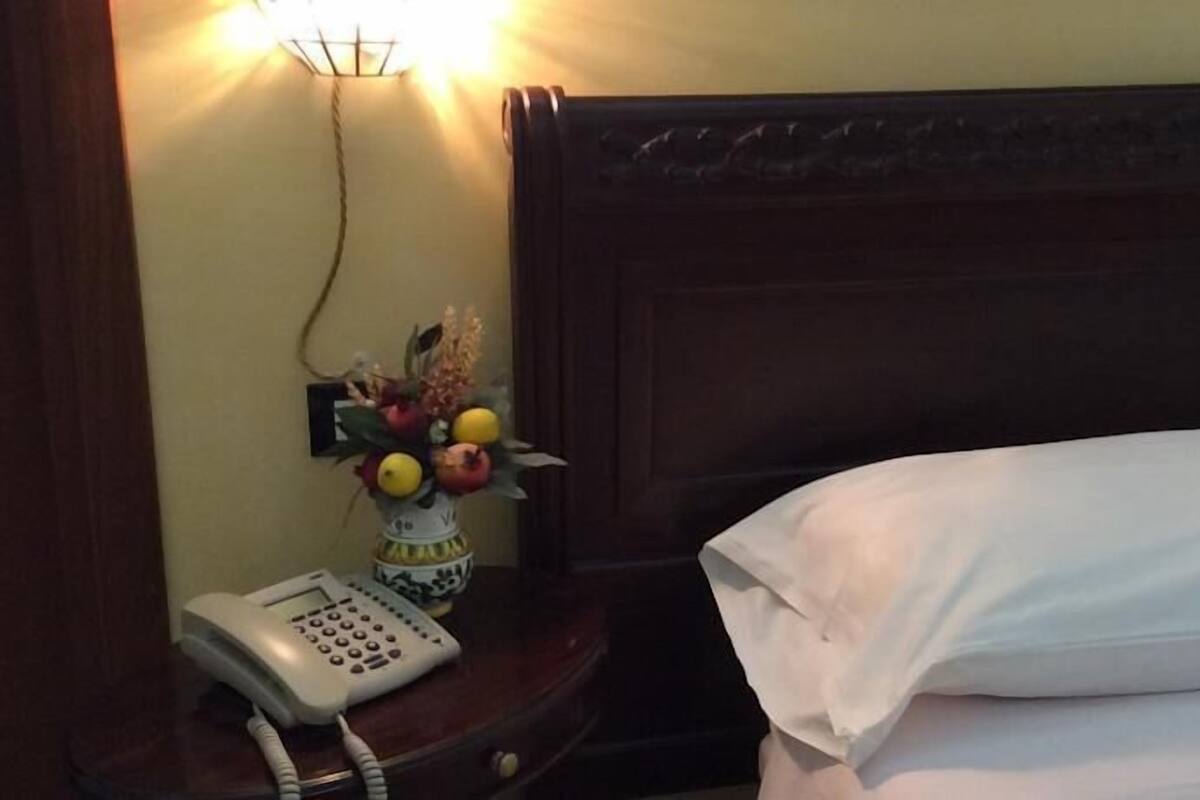 Habitación de hotel con cabecera de madera, mesita de noche con teléfono y un jarrón de flores, y almohada blanca en la cama.