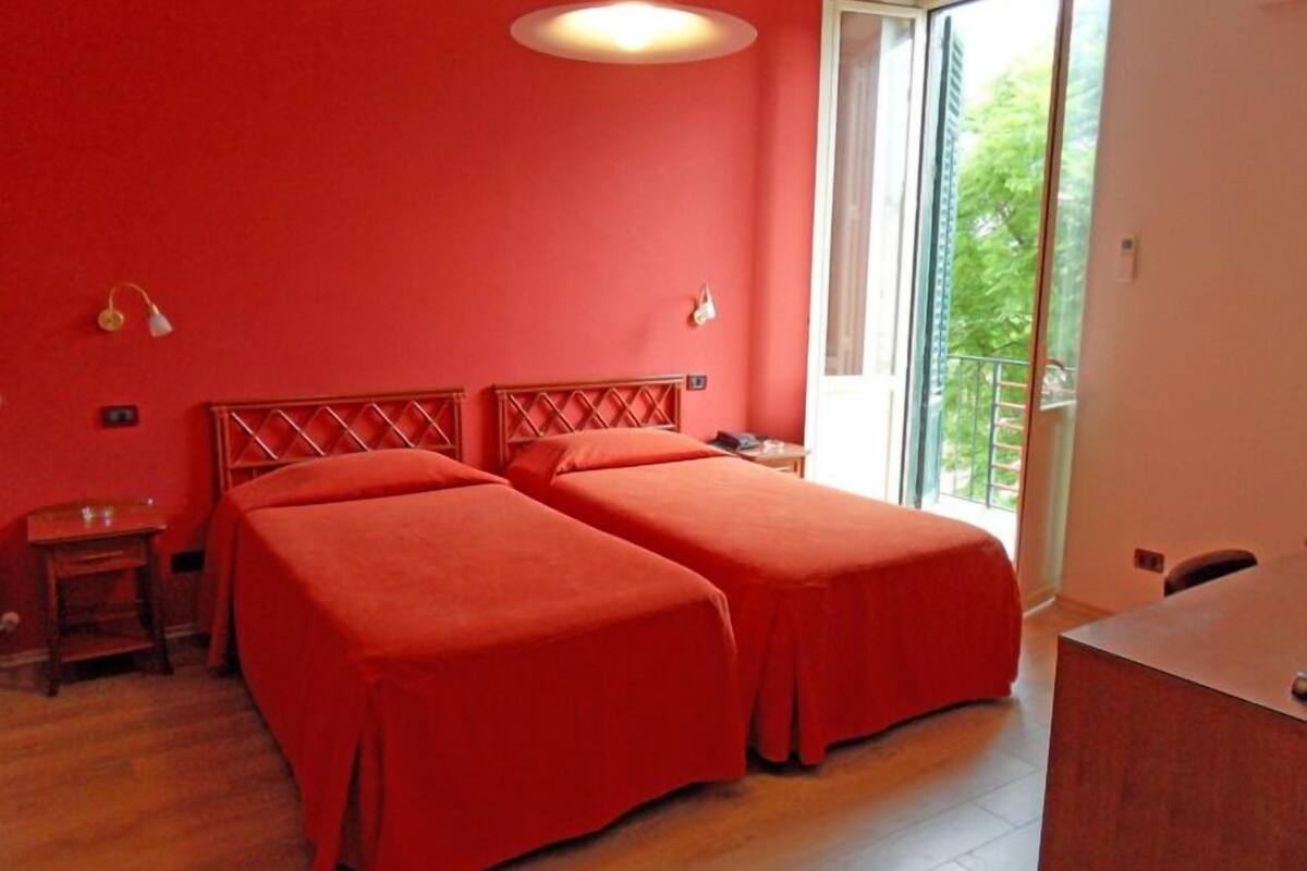 A room with two beds, a wooden floor, a balcony, and a large window.