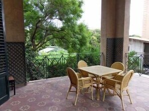 Uma área de estar coberta ao ar livre, com mesa e cadeiras, rodeada por um chão estampado e um corrimão de metal.