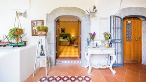 A hallway with a tiled floor, a white table with a vase of flowers, a hanging plant, and a door with a metal gate. - Orchid Corner (Meta)