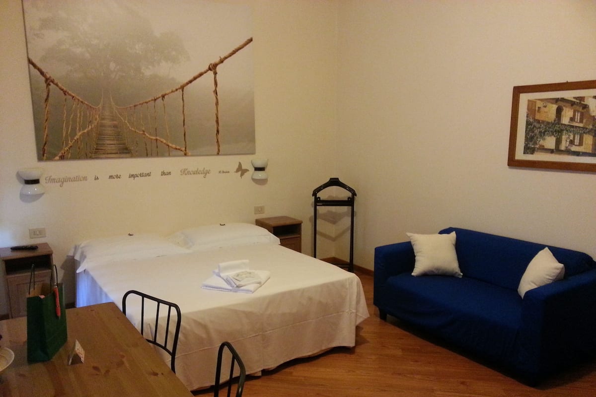 A room with a bed, a blue sofa, a wooden table, and a painting of a bridge.