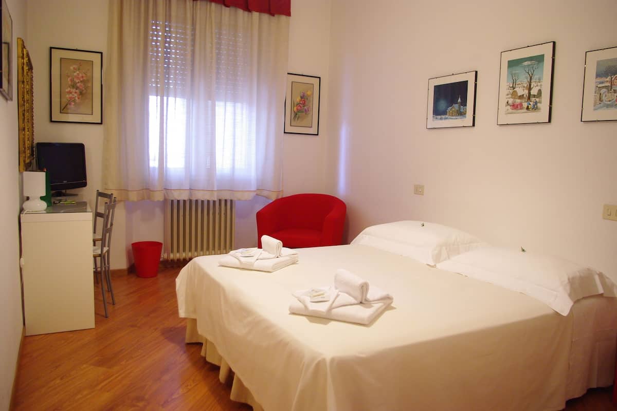 A hotel room with a bed, a desk with a computer, a chair, and a red armchair. There are pictures on the wall and a window with curtains.
