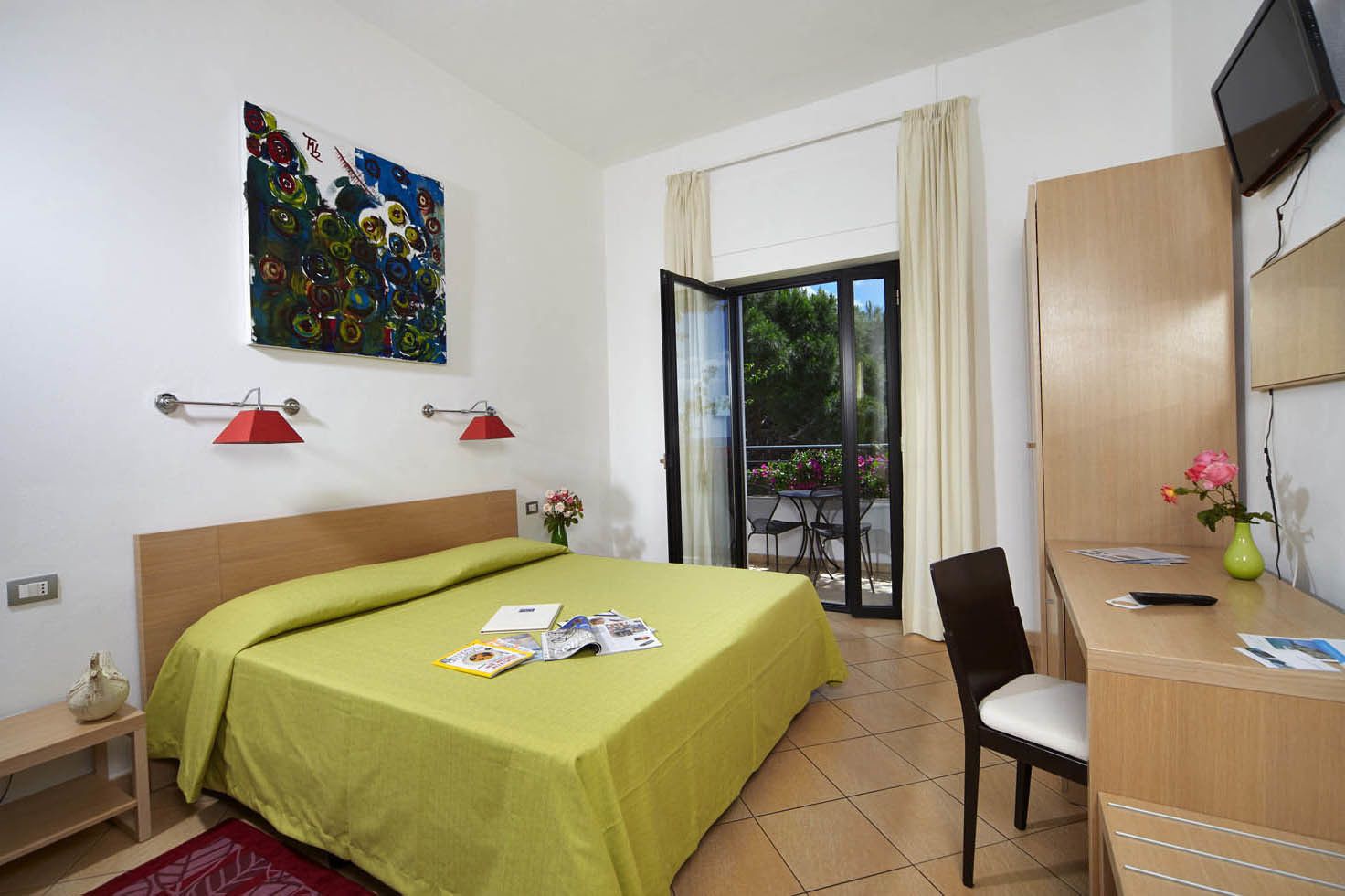 Habitación de hotel con cama, escritorio, silla, televisor y balcón con mesa y sillas.