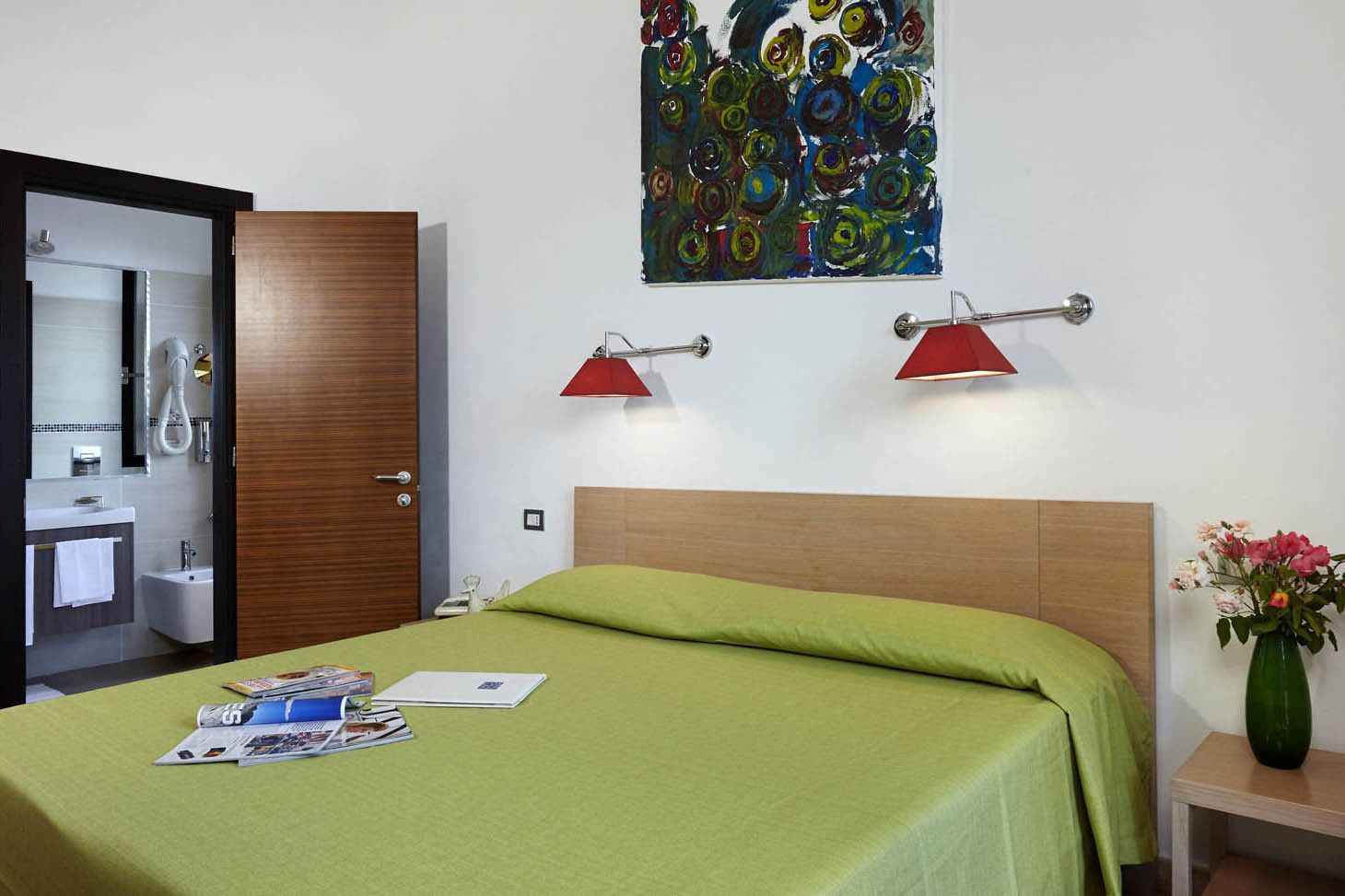 Camera d'albergo con un letto, un armadio in legno, bagno con specchio, lavandino e doccia. C'è un quadro appeso al muro e un vaso con fiori sul comodino.