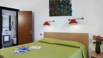 Camera d'albergo con un letto, un armadio in legno, bagno con specchio, lavandino e doccia. C'è un quadro appeso al muro e un vaso con fiori sul comodino.