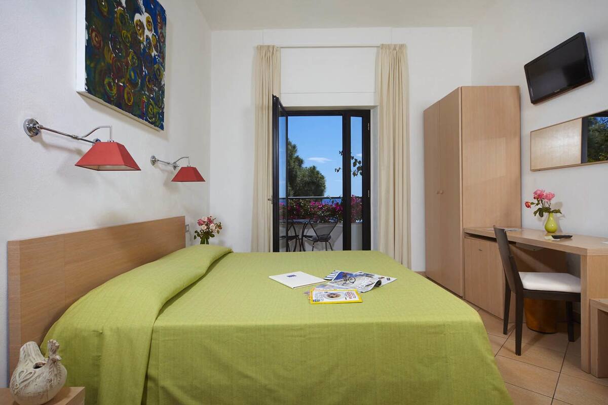 Hotellihuone, jossa on sänky, työpöytä, tuoli, televisio ja parveke, jolta on näkymä.