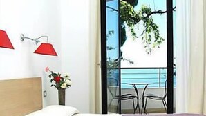 Habitació d'hotel amb un llit, una tauleta de nit amb un vas de flors, un balcó amb taula i cadires, i vistes al mar.