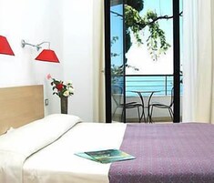 Habitación de hotel con cama, mesita de noche con un jarrón de flores, balcón con mesa y sillas, y vista al mar.
