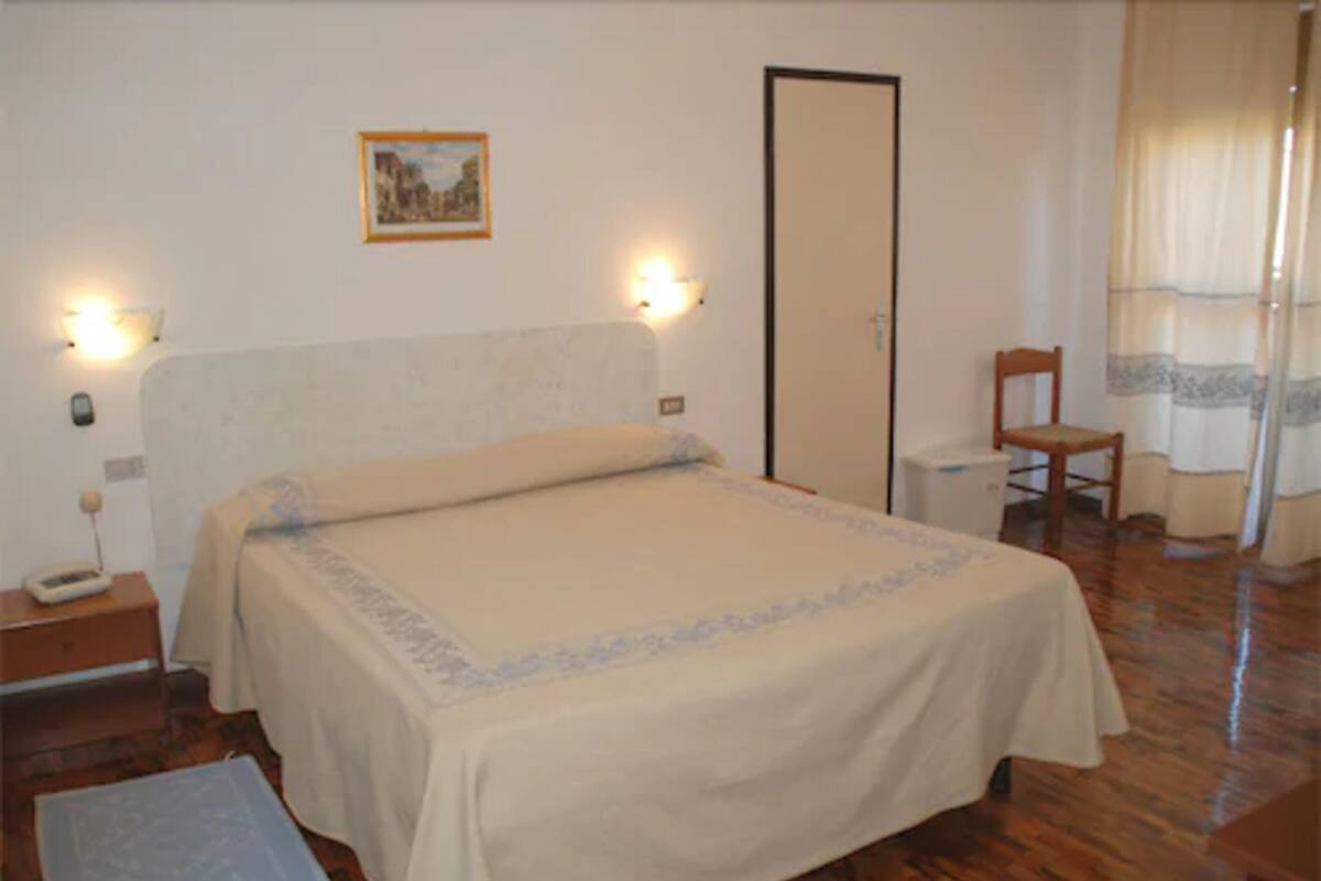 Un dormitorio con cama, mesitas de noche, una silla, un armario y un cuadro en la pared.