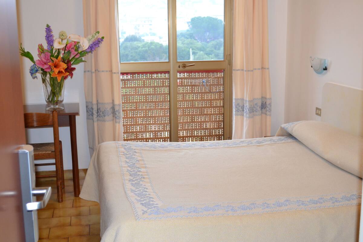 Un dormitorio con cama, una silla, un jarrón con flores y ventana con vistas a árboles.