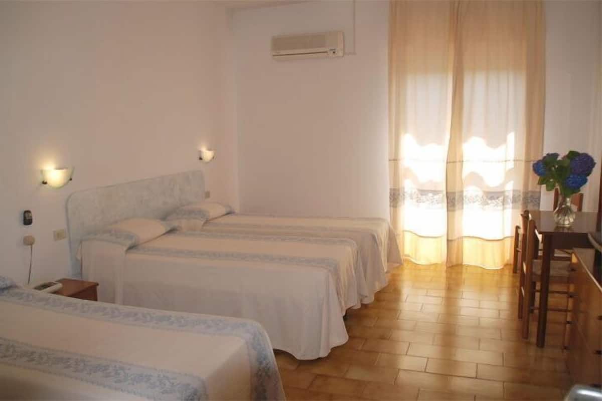 Habitación de hotel con dos camas individuales, un escritorio y una silla. Se ve una ventana con cortinas, un equipo de aire acondicionado en la pared y una pequeña mesa con un florero.