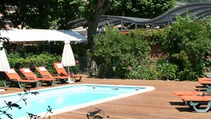 Una piscina con sillas de descanso y sombrillas, rodeada de vegetación y una terraza de madera.