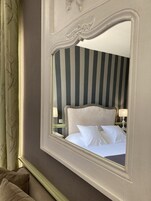 Una habitación de hotel con un espejo grande que refleja una cama con ropa blanca y una pared a rayas al fondo.