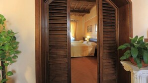 Une pièce avec une porte en bois donnant sur un lit doté d’une tête de lit, une table de chevet avec une lampe et une plante en pot.