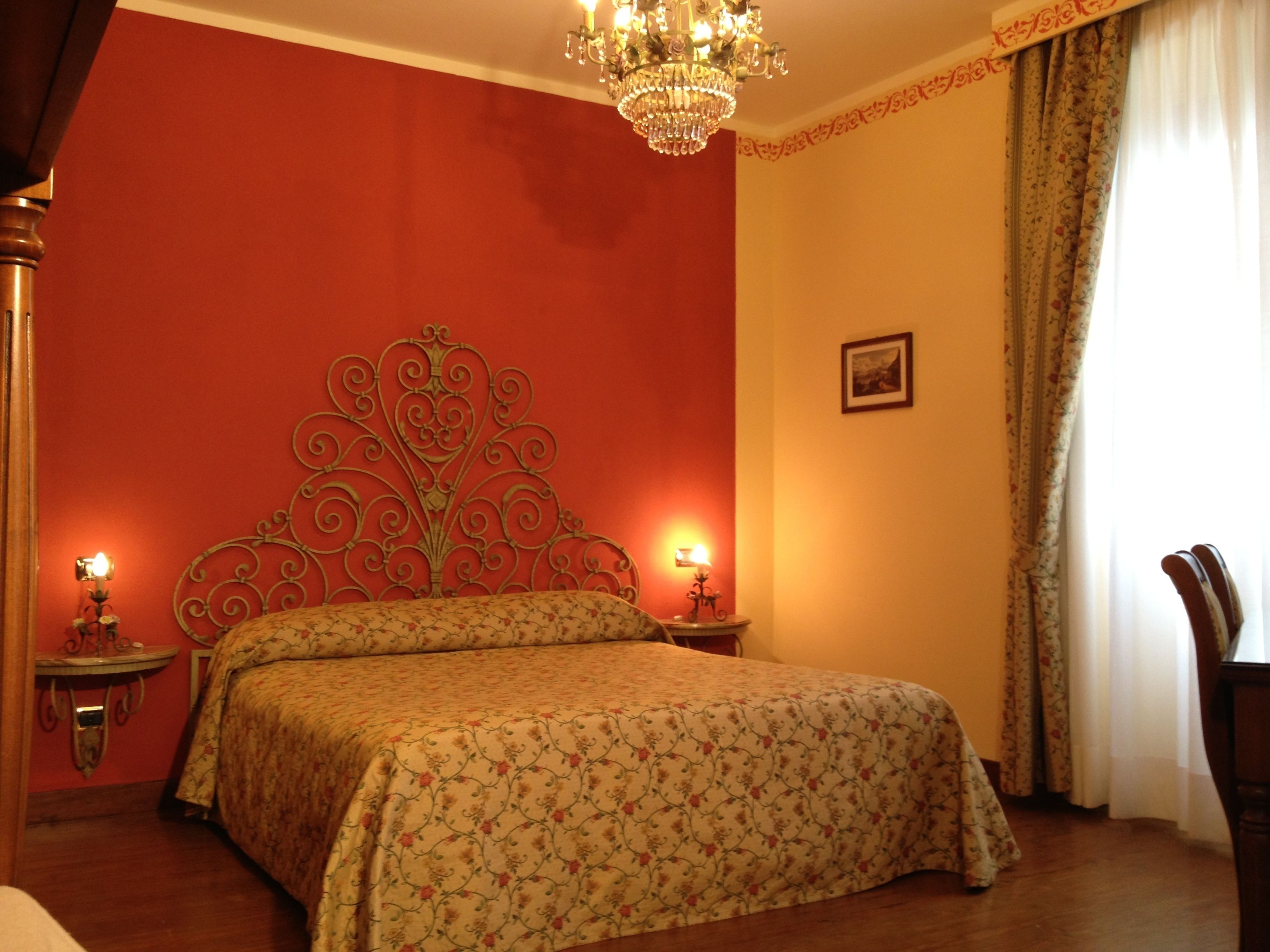 A bedroom with a large bed, two bedside tables with lamps, a chandelier, and a window with curtains.