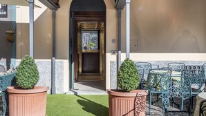 Entrada do Hotel du Lac, com uma placa acima da porta, uma pequena área de jardim com vasos de plantas e mesas externas com cadeiras de metal.