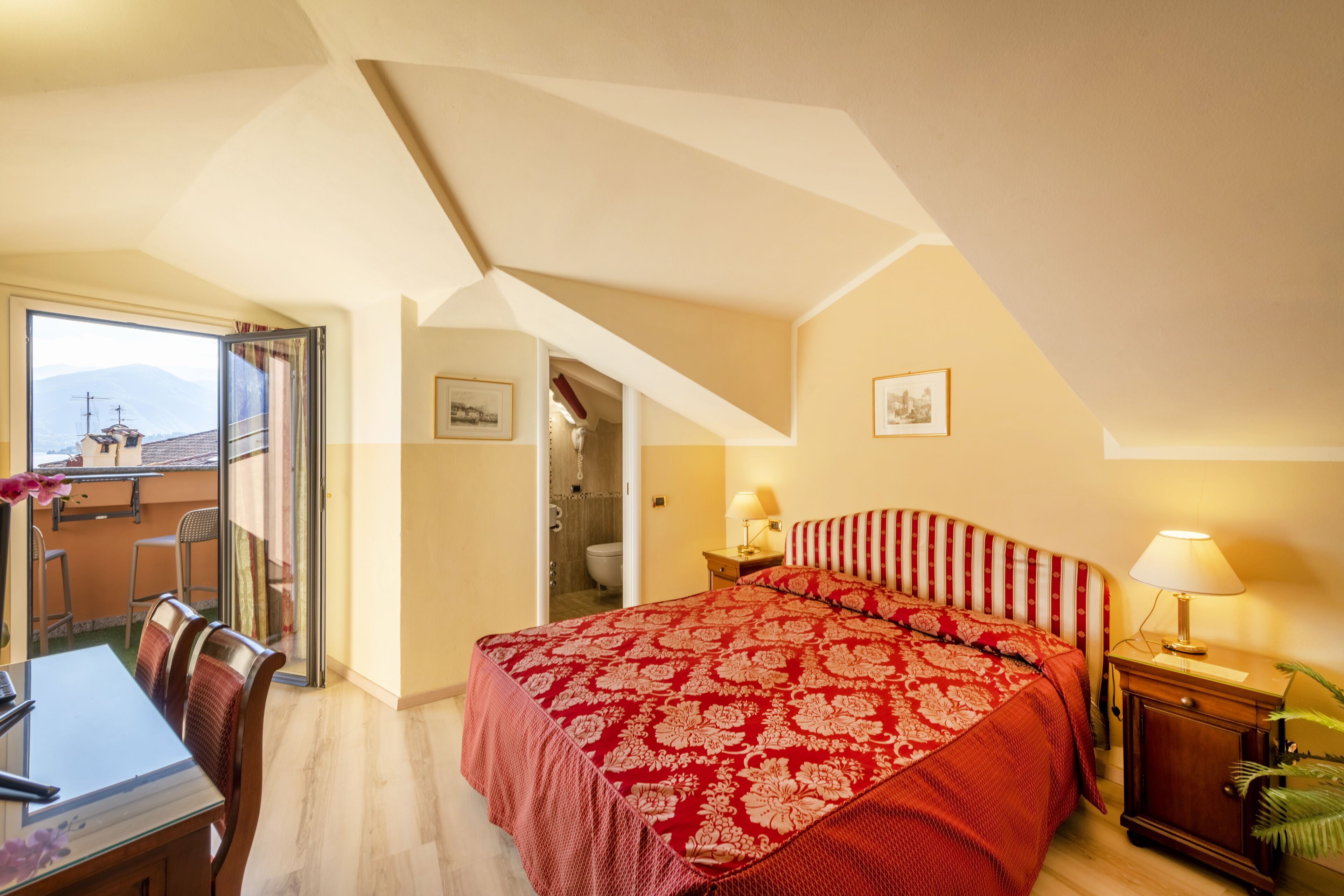 A bedroom with a large bed, a desk, and a chair. There is a window with a view of mountains.