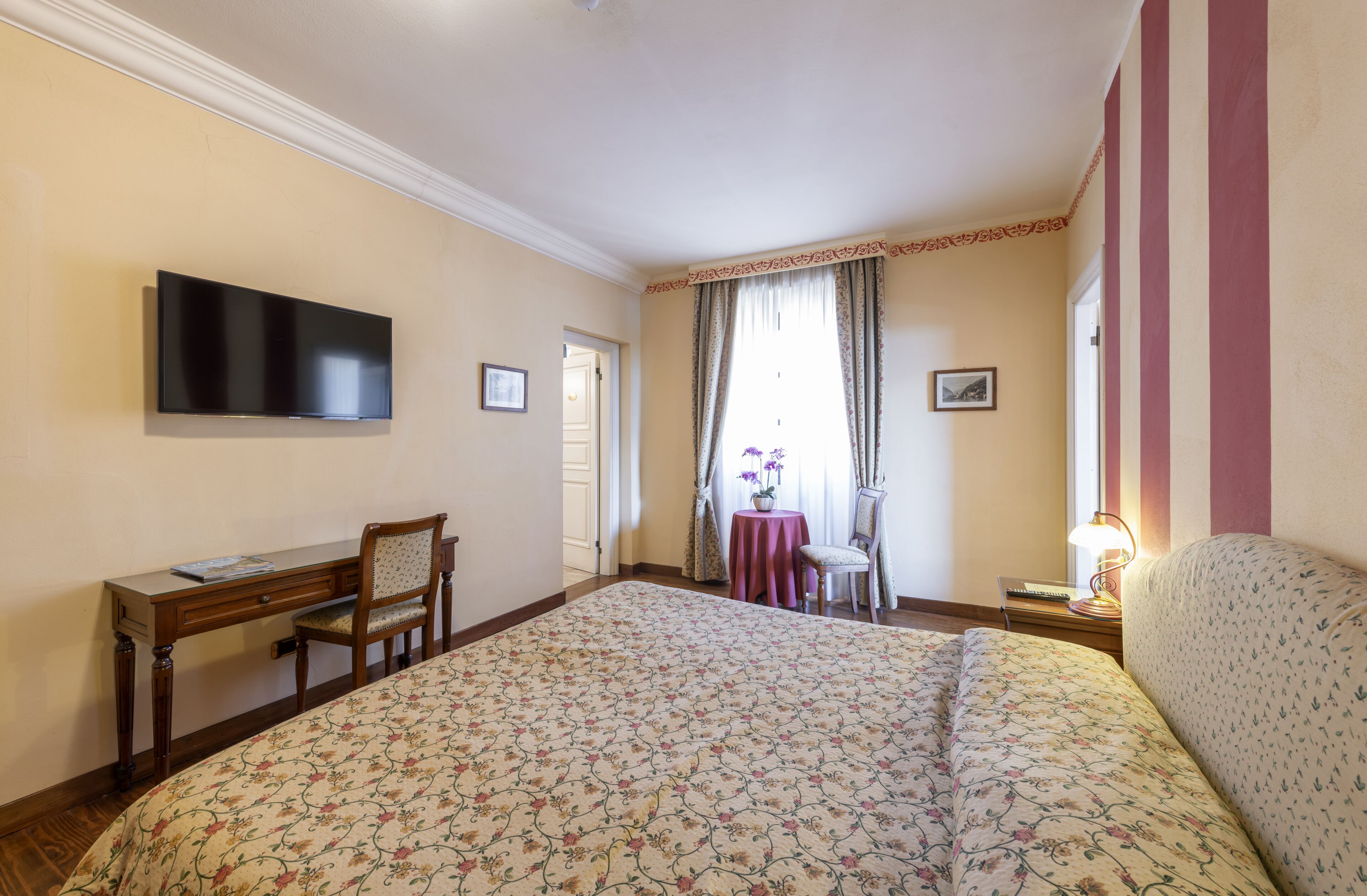 A hotel room with a large bed, a desk with a chair, a television mounted on the wall, a small table with a chair, and a window with curtains.