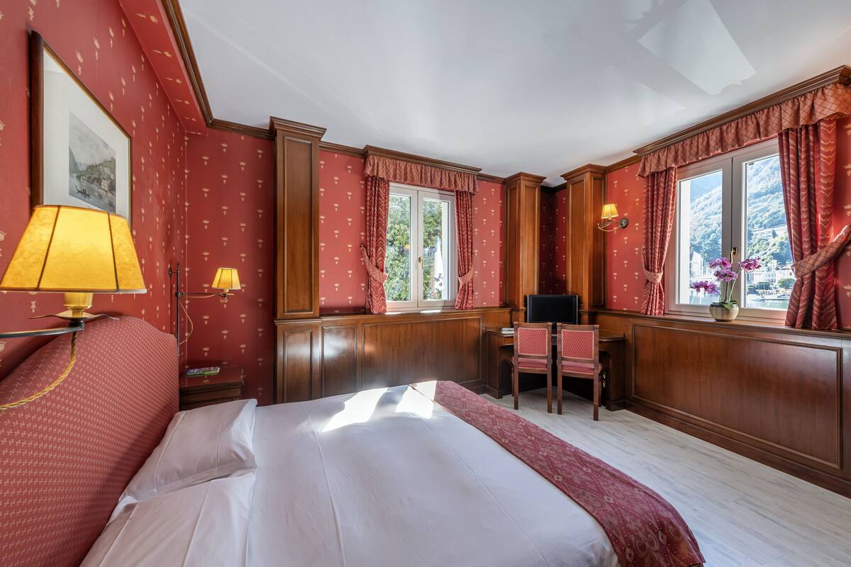 Habitación de hotel con una cama grande, dos ventanas con cortinas, un escritorio con una silla y una lámpara.