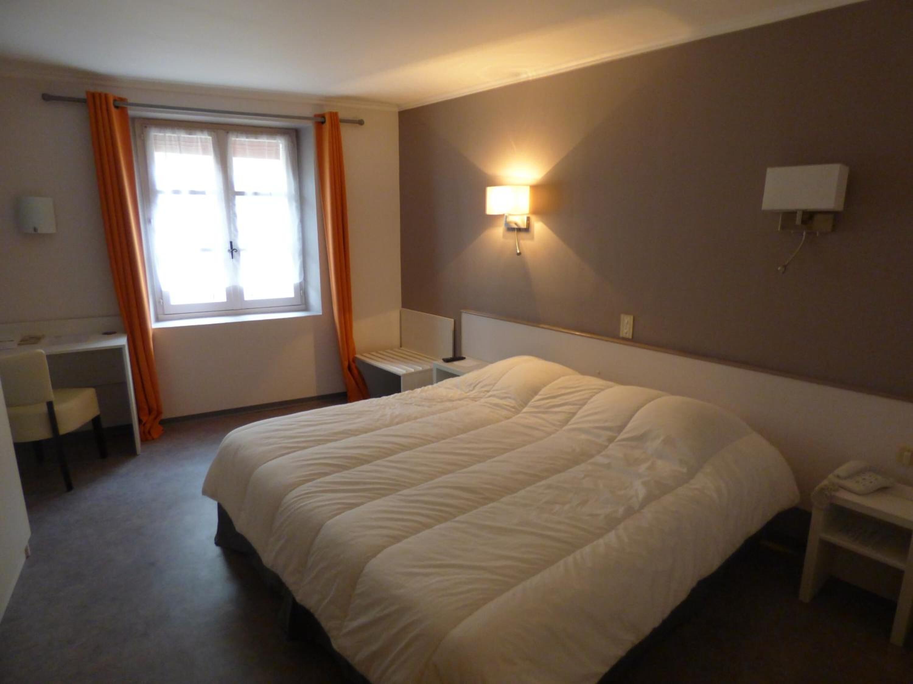 Habitación de hotel con una cama grande, un escritorio y una silla. Se ve una ventana con cortinas, una lámpara de pared y un teléfono en una mesita de noche.