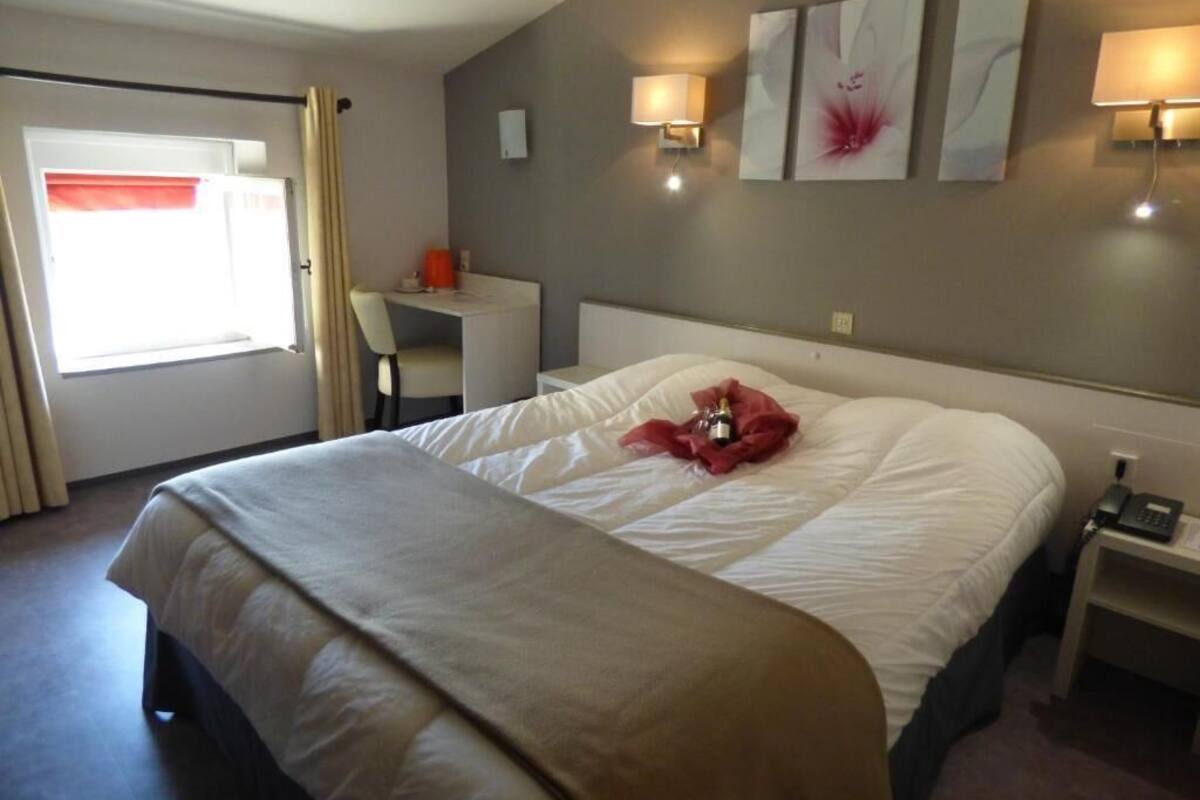A hotel room with a large bed, a desk, and a chair. There are wall-mounted lamps and a window with curtains.
