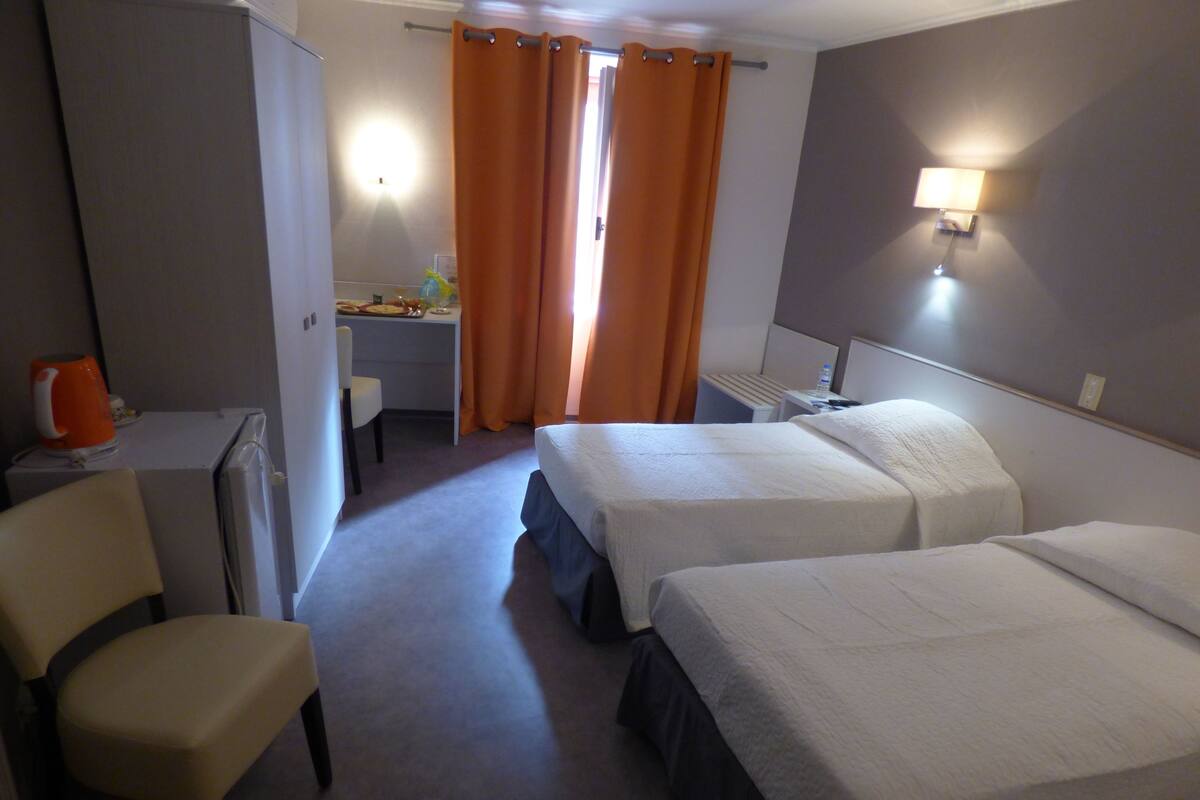 A hotel room with two beds, a desk, a chair, and a bathroom visible through a curtain.