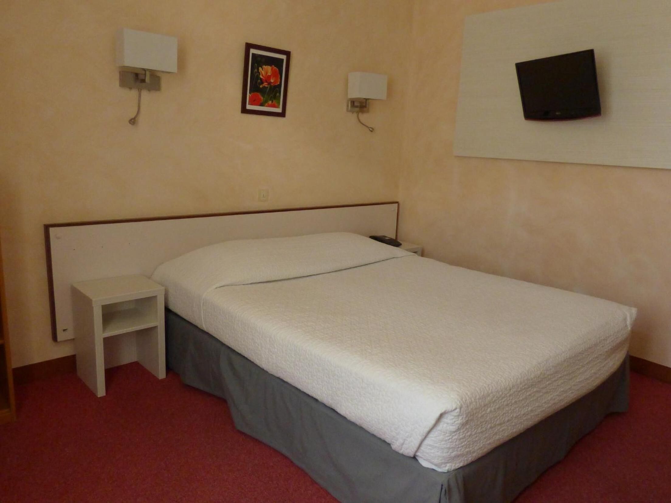 A single bed with a grey skirt, a white nightstand, a framed picture on the wall, and a flat-screen TV mounted above the bed.