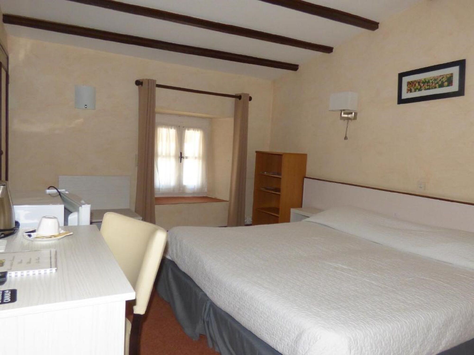 A hotel room with a bed, a desk with a chair, a window with curtains, a bookshelf, and a wall-mounted lamp.