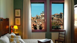 Un dormitorio con cama, una silla, ventana con vistas a edificios, lámpara y cuadros en la pared.