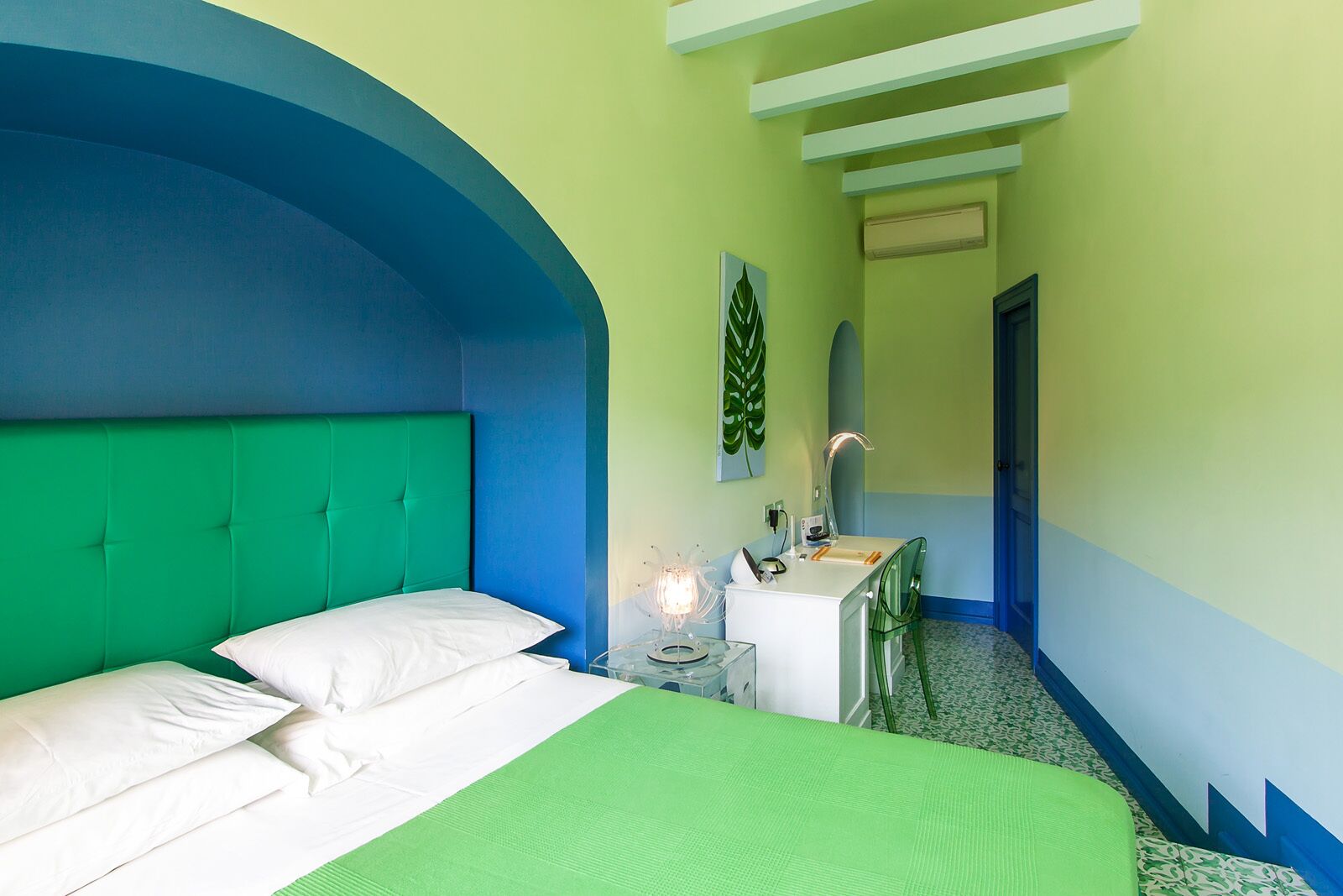 A hotel room with a bed, a bedside table, a mirror, and a green wall.