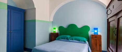 A bedroom with a bed, green headboard, two bedside tables with lamps, a blue wardrobe, and patterned floor tiles.
