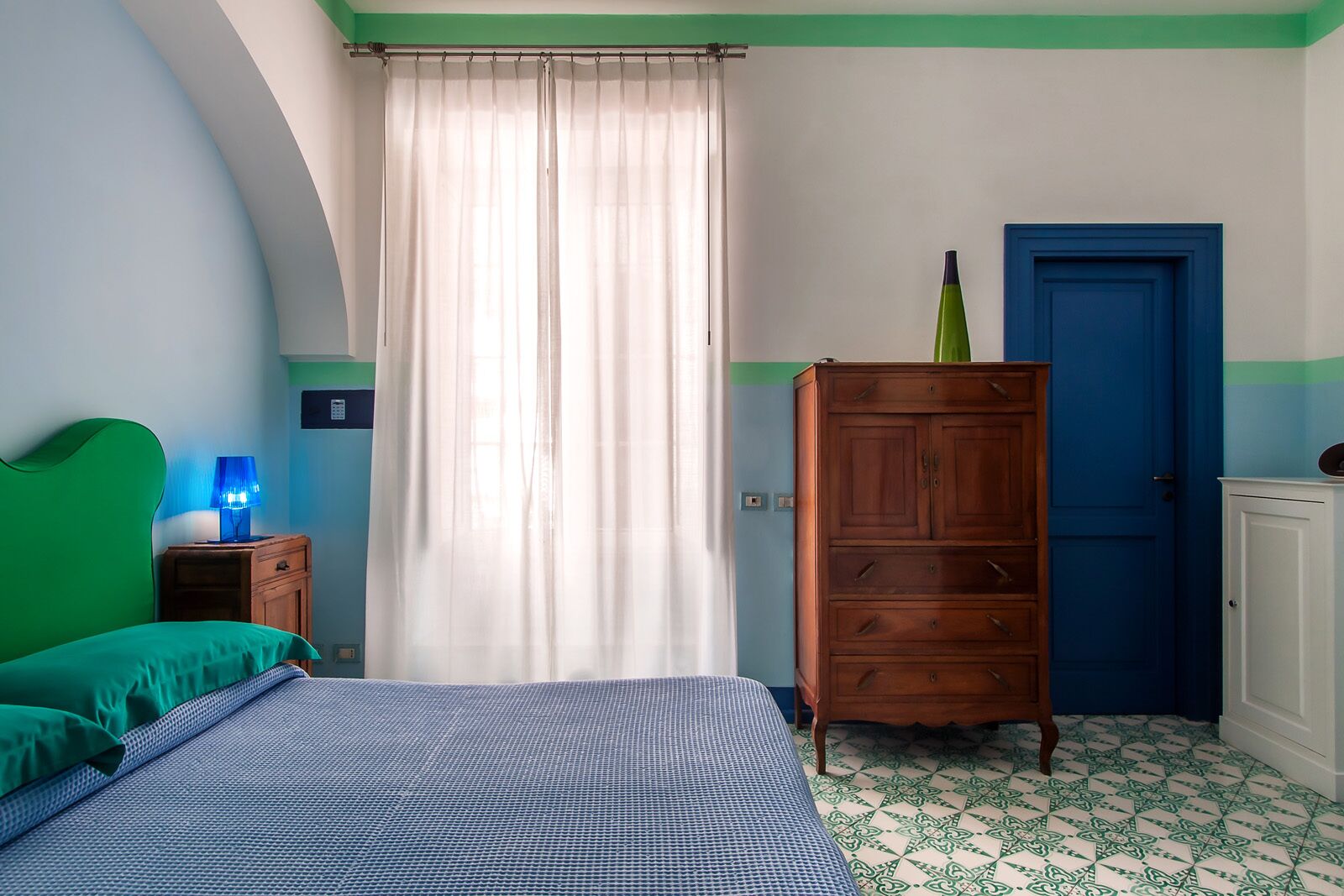 A bedroom with a bed, a nightstand, a dresser, a blue door, and a window with curtains.