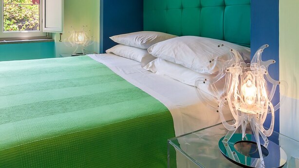 A bedroom with a bed, a glass side table, a lamp, and a window with a view of greenery. - Casa Astarita (Sorrento)