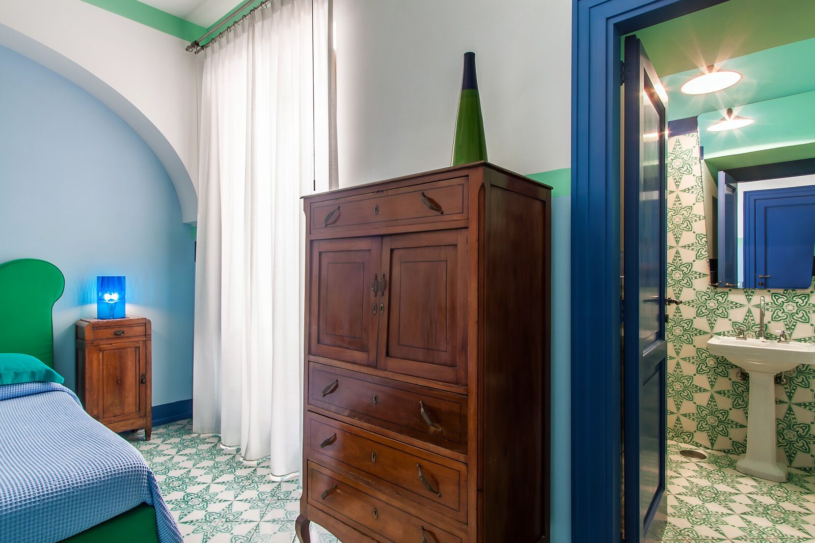 A bedroom with a bed, a wooden dresser, a nightstand with a lamp, and a bathroom with a sink and mirror.